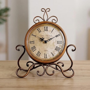 May include: A vintage-style, bronze-colored clock with a round face and Roman numerals. The clock features a decorative stand with scrolled details and the text "Insto Clock & C 1845".