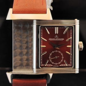 May include: A rectangular silver-tone wristwatch with a deep burgundy dial, featuring the text Jaeger-LeCoultre and Swiss Made. The watch has a sub-dial for seconds, silver-tone hour markers, and a textured metal side panel, attached to a reddish-brown strap.
