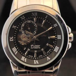 May include: A silver-tone stainless steel analogue watch with a black dial, Roman numeral hour markers, and a metal link bracelet. The watch face features a visible open-heart movement, a 24-hour sub-dial, and the text Premier AUTOMATIC MADE IN JAPAN.