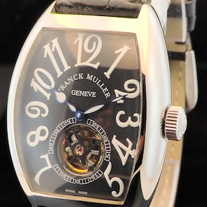 May include: A Franck Muller Geneve wristwatch with a tonneau-shaped silver case and a black leather strap. The black dial features large white Arabic numerals, a visible tourbillon movement at the bottom, and blue hands. Swiss Made is printed.