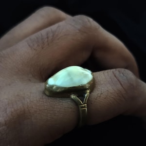 May include: A gold-toned ring featuring a large, oval-shaped, off-white stone. The ring has a simple band and a decorative setting that holds the stone. The ring is worn on a finger.