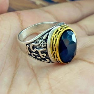 May include: Silver ring with a large, oval, dark blue gemstone. The ring has intricate detailing on the band and a gold-colored bezel around the stone. The ring is displayed on a light-colored surface.