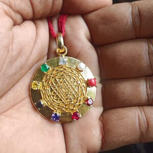 Könnte beinhalten: Goldfarbener Anhänger mit geometrischem Design und eingelegten Edelsteinen in verschiedenen Farben. Der Anhänger ist rund und hängt an einer roten Kordel. Die Edelsteine umfassen Grün, Gelb, Blau und Rot.