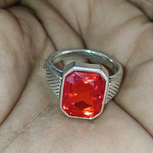 May include: A silver ring featuring a large, rectangular, faceted red gemstone. The ring has a textured design on the sides of the band, adding detail to the jewelry. The gemstone is the focal point, catching the light.