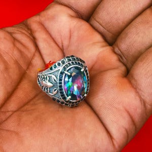 May include: A silver ring with an oval, iridescent gemstone. The ring is set with small, dark blue stones around the main stone and along the band. The ring is held in a hand against a red background.