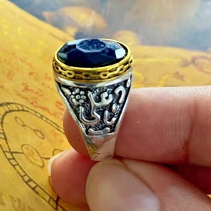 May include: A silver ring with a large, dark blue gemstone. The ring has a gold-colored band around the gemstone and ornate detailing on the sides, including what appears to be Arabic script. The ring is held up by a person's fingers.
