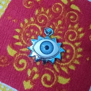 May include: A silver-toned pendant featuring a blue eye design with a white border, set within a sunburst frame. The pendant is displayed on a red patterned fabric.
