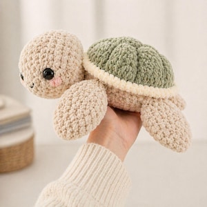 Könnte beinhalten: Ein gehäkelter Schildkröten-Plüsch in Beige und Grün. Die Schildkröte hat einen beigen Kopf und Gliedmaßen, einen grünen Panzer und einen cremefarbenen Rand. Sie hat schwarze Augen und rosige Wangen und wirkt dadurch freundlich.