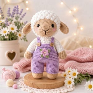 May include: A handmade crochet lamb doll with a white head and body, wearing purple overalls with a flower applique. The lamb has tan arms and legs, and black eyes. The doll is displayed on a wooden stand with flowers in the background.