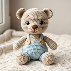 Könnte beinhalten: Ein gehäkelter Teddybär mit hellbraunem Körper und Kopf, der eine hellblaue Latzhose mit Holzknöpfen trägt. Der Bär hat schwarze Augen und eine braune Nase und sitzt auf einer weißen, strukturierten Decke. Handgefertigtes Spielzeug.