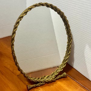 May include: A round mirror featuring a metallic gold frame with a textured, braided rope design. The mirror is supported by a matching gold-toned base that extends outward with two small, flared feet at the bottom of the frame.