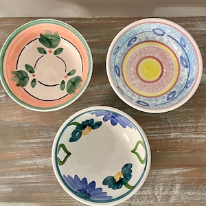 May include: Three decorative ceramic bowls with colorful floral designs. One bowl has a pink and green floral pattern, another has a blue and yellow design, and the third features blue flowers. The bowls are set on a light brown wooden surface.