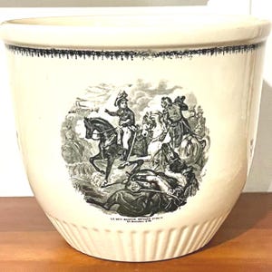 May include: A cream-coloured ceramic planter featuring a black transferware print of a historical battle scene with figures on horseback. The rim is decorated with a black scalloped border, and the base has vertical fluting. Text reads LE GEO MASSEM DEVANT ZURICH 25 Decembre 1799.