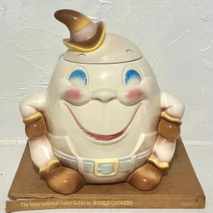 May include: A ceramic Humpty Dumpty cookie jar featuring a cream-colored egg-shaped body, a brown hat, blue eyes, and pink cheeks. The character wears a light-colored belt with a yellow buckle and brown gloves, resting on a brown book.