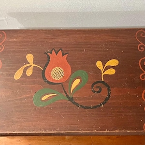 May include: A rectangular wooden box with a dark brown stained finish. The lid features a hand-painted floral motif in the center, showcasing a red tulip-like flower with a yellow grid pattern, green leaves, and yellow accents, framed by orange scrollwork.