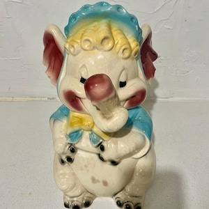 Peut inclure: Une figurine d'éléphant en céramique au corps crème, portant un bonnet bleu et un nœud jaune, avec des touches de rose sur les oreilles et les joues. L'éléphant a la trompe recourbée vers le haut, les yeux fermés et des cheveux texturés.