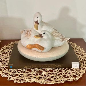 Könnte beinhalten: Eine Keramikfigur von zwei weißen Enten, die auf einer strukturierten, kreisförmigen Basis ruhen. Die Enten haben braune Schnäbel und detaillierte Federzeichnungen. Die Figur steht auf einem dunkelbraunen Buch mit dem Titel Aesop's Fables.