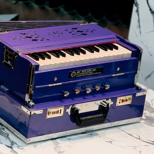May include: A blue and purple harmonium with a keyboard and bellows. The instrument has a handle and is sitting on a round table. The text "JC MUSICAL OFFICIAL" is visible in the bottom left corner.