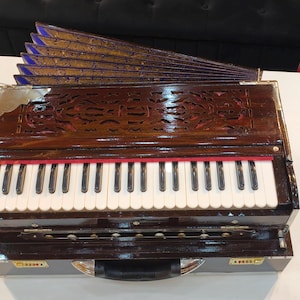 May include: A brown and gold harmonium with a keyboard of white and black keys. The instrument has a decorative carved wooden top and a fan-shaped structure at the top. The harmonium sits on a white surface.