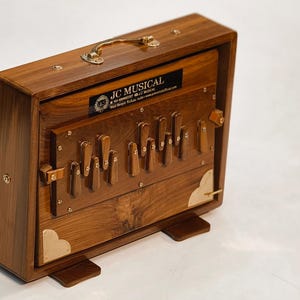 May include: A wooden harmonium, rectangular in shape, with a handle and brass details. The instrument has vertical keys and a label reading "JC MUSICAL". It rests on two small wooden feet. The harmonium is made of wood.
