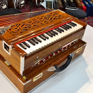 May include: A wooden harmonium with a keyboard and bellows. The instrument has a brown finish with decorative carvings on the top. The keyboard has black and white keys. A black handle is on the front.