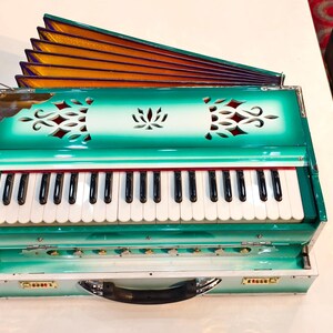 May include: A teal and white harmonium with a keyboard and a fan-shaped bellows. The instrument has decorative cutouts and a handle. The bellows are gold and purple. The text "JC MUSICAL OFFICIAL" is visible.