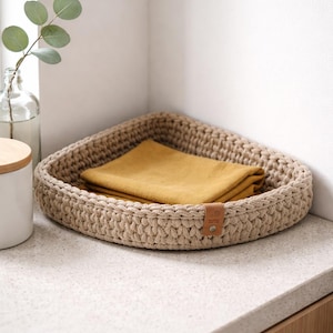 Könnte beinhalten: Ein beiger, dreieckiger Häkelkorb mit einem senfgelben, gefalteten Tuch im Inneren. Der Korb hat ein Lederetikett. Der Korb steht auf einer hellen Arbeitsplatte, mit einer Glasflasche und einem weißen Glas im Hintergrund.