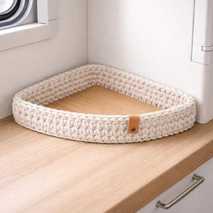 Könnte beinhalten: Ein cremefarbener, dreieckiger Häkelkorb mit braunem Lederakzent. Der Korb steht auf einer hellen Holzoberfläche und eignet sich ideal zum Ordnen kleiner Gegenstände. Das Design ist schlicht und funktional, passend für verschiedene Einrichtungsstile.