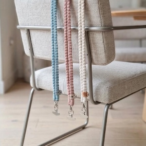 Könnte beinhalten: Drei Makramee-Handy-Lanyards in Pastellfarben hängen an einem Stuhl. Die Lanyards sind hellblau, rosa und cremefarben, jeweils mit einem silberfarbenen Verschluss. Der Stuhl hat einen grauen Stoffbezug und ein silbernes Metallgestell.