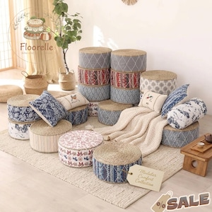 May include: A selection of round floor cushions and throw pillows. The cushions have woven straw tops and patterned fabric sides. Pillows feature floral and bird designs. A cream-coloured blanket and a small wooden table are also present.