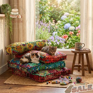 May include: A pile of vibrant, patterned floor cushions with pom-pom edging. Two kittens are sitting on the cushions. A small wooden stool holds a mug that says "Cat Nap" and glasses. A window with a garden view is in the background.