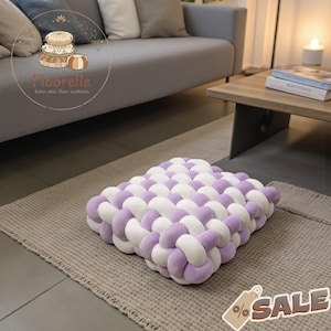 Può includere: Un cuscino da pavimento quadrato con un design intrecciato in lavanda e bianco. Il cuscino è realizzato in materiale morbido e poggia su un tappeto di colore neutro. Il cuscino fa parte della collezione "Floorelle boho-chic floor cushions".