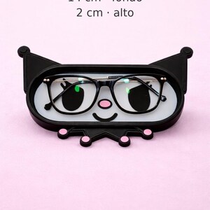 May include: A black eyeglasses holder shaped like a cartoon character's face, with a white interior and pink accents. The holder contains a pair of black-framed glasses. Dimensions are 8.3 inches wide, 5.5 inches deep, and 0.8 inches high.