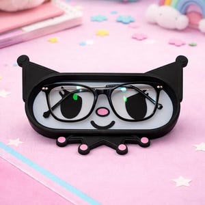 May include: Black eyeglasses rest in a black and white cat-shaped glasses holder with pink accents. The holder has a smiling face design and is on a pink surface with decorative stars and a rainbow.