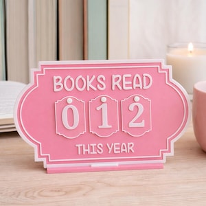 Puede incluir: Un letrero decorativo rosa y blanco con las palabras "BOOKS READ THIS YEAR" y los números "012". El letrero está diseñado para llevar un registro de los libros leídos. Se muestra sobre una superficie de madera.