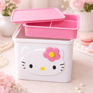 Puede incluir: Caja de almacenamiento blanca con tapa rosa y bandeja extraíble. La caja presenta un diseño de Hello Kitty con una flor rosa y centro amarillo. La caja está sobre una superficie clara con flores rosas en el fondo.