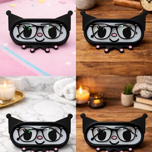 May include: Black eyeglass holder with a cartoon face design. The holder has a white interior and a black exterior with a smiling face, glasses, and pink accents. The holder is designed to hold a pair of eyeglasses.