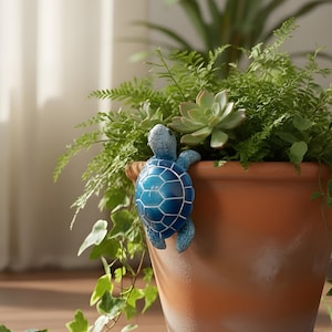 Puede incluir: Una maceta de terracota llena de varias plantas verdes, incluyendo helechos y suculentas. Una tortuga decorativa azul está sujeta al borde de la maceta. La escena está ambientada en interiores con luz natural y suelo de madera.