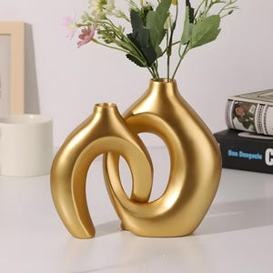 May include: A gold-coloured ceramic vase featuring two interconnected, curved, hollow circular forms with two distinct openings at the top. The vase holds a small bouquet of artificial white, yellow, and pink flowers with green leaves.