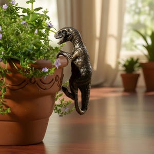 May include: A bronze-colored dinosaur figurine clinging to the edge of a terracotta flower pot filled with green plants and small purple flowers. The dinosaur has detailed features and is positioned as if climbing.