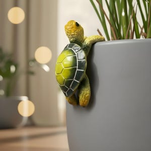 Puede incluir: Una figura decorativa de tortuga en tonos amarillos, verdes y negros, aferrada al lateral de una maceta gris. La tortuga tiene un caparazón texturizado y está colocada cerca de tallos de plantas verdes.