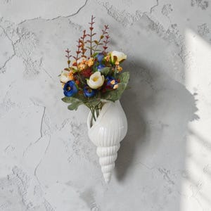 May include: A handmade, wooden sign with the text "Welcome" in a handwritten style. The sign is painted white with a distressed finish, and a small, green plant is attached to the bottom. The sign is designed for indoor or outdoor use.