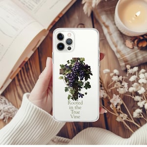 Può includere: Custodia trasparente per telefono con un design a croce in legno, adornata da uva viola e foglie verdi. Il testo "Rooted in the True Vine" è stampato sotto. La custodia è tenuta su una superficie di legno.