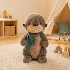 Könnte beinhalten: Ein kuscheliges Otter-Spielzeug mit grauem Körper, brauner Nase und einem teal-farbenen Schal. Das Stofftier sitzt auf einem cremefarbenen Teppich. Im Hintergrund sind Holzspielzeuge und ein kleines Tipi zu sehen.