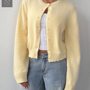 May include: A pale yellow cardigan with a round neckline and button-down front. The long-sleeved sweater is paired with light blue jeans. The cardigan has ribbed cuffs and hem.