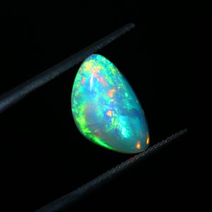 May include: An oval opal gemstone, held by tweezers against a black backdrop. The opal exhibits a captivating play of colour, with flashes of green, blue, orange, and yellow. The gemstone's surface is smooth and polished, reflecting light.