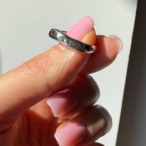 Puede incluir: Un anillo plateado con la palabra "SHINE" grabada. El anillo tiene pequeñas piedras preciosas rojas y verdes. El anillo lo sostiene una persona con esmalte de uñas rosa.