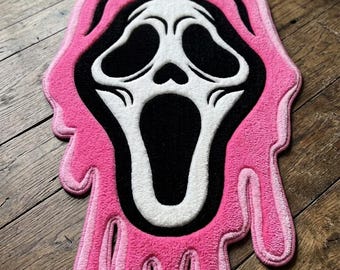 May include: A hand-tufted rug featuring a white and black ghost face mask design surrounded by a bright pink melting effect. The rug has a rectangular brown leather tag at the bottom with the text HAND-TUFTED PROPER printed in black ink.
