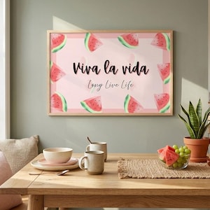 May include: A framed print with a pink background and watermelon slices. The text "Viva la vida, Long Live Life" is written in black script. The print hangs above a wooden table with a bowl of fruit and a potted plant.