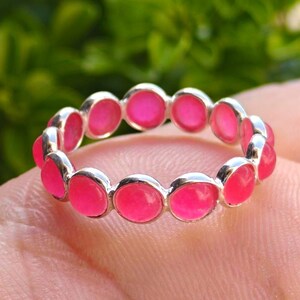 Könnte beinhalten: Ein silberner Ring mit einem Band aus leuchtend rosa Edelsteinen. Der Ring hat eine Reihe von runden, bündig gefassten Steinen, die ein durchgehendes Muster bilden. Die Edelsteine sind ein helles, sattes Rosa, das mit der silbernen Fassung kontrastiert.